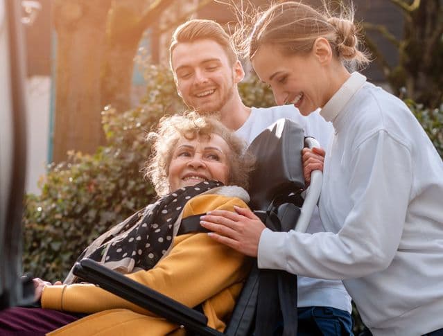 Elderly woman in wheelchair receiving assistance from two young people